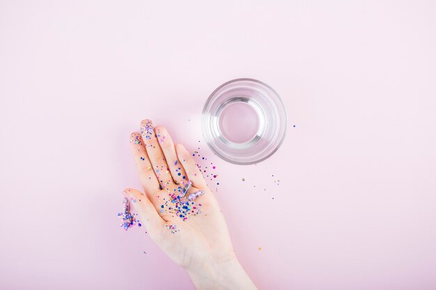 Paillettes e glitter capsula sulla mano umana vicino al bicchiere d&#39;acqua su sfondo rosa