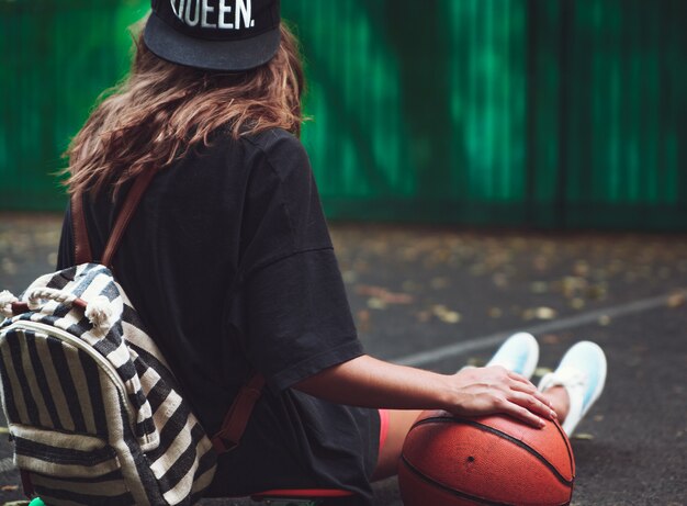 Palla di pallacanestro della foto del primo piano con la ragazza che si siede sulla shortboard arancio di plastica del penny sull'asfalto