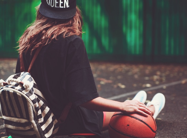 Palla di pallacanestro della foto del primo piano con la ragazza che si siede sulla shortboard arancio di plastica del penny sull'asfalto