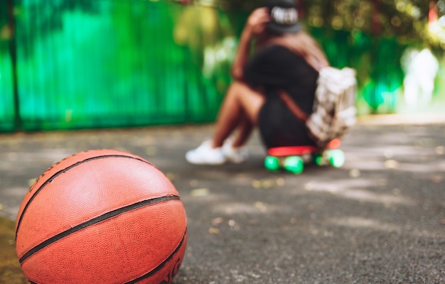 Palla di pallacanestro della foto del primo piano con la ragazza che si siede sulla shortboard arancio di plastica del penny sull'asfalto