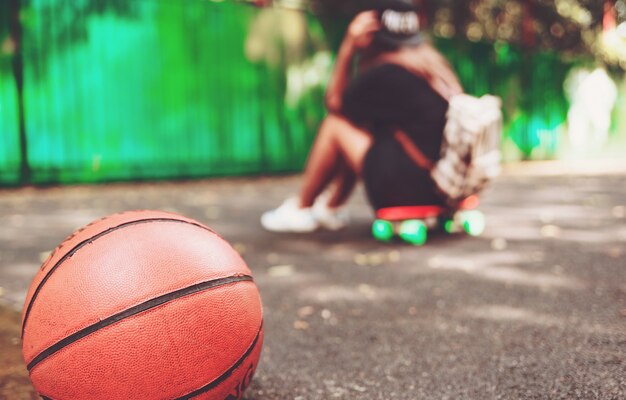 Palla di pallacanestro della foto del primo piano con la ragazza che si siede sulla shortboard arancio di plastica del penny sull'asfalto