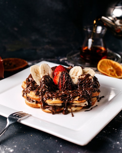 Pancake di choco di vista frontale con banane e fragole rosse all'interno del piatto bianco sulla scrivania luminosa