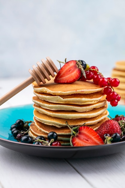 Pancake di vista frontale con fragole ribes nero e rosso su un piatto con un bastone per miele