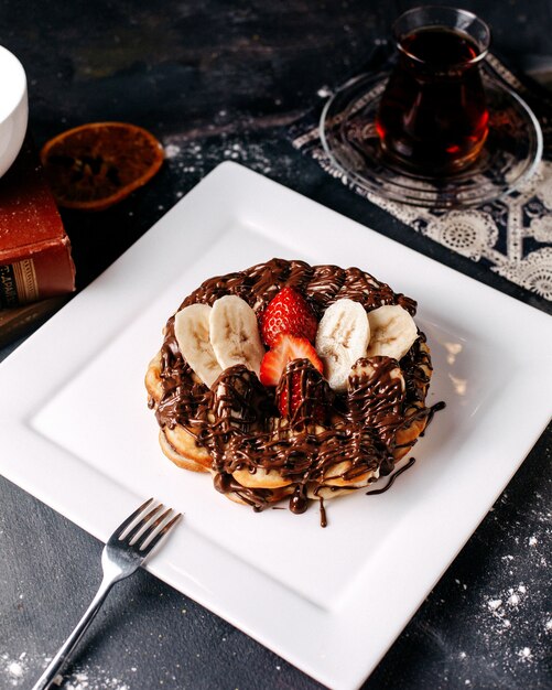 Pancake squisito di vista frontale choco insieme alle fragole e alle banane rosse affettate dentro il piatto bianco sul pavimento grigio