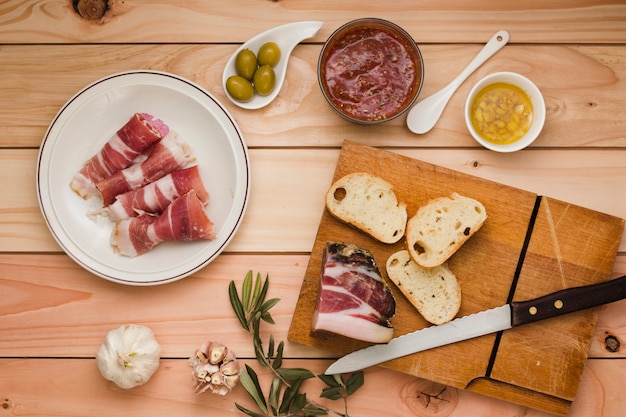 Pancetta cruda; salsa di salsa; olive; pane tostato; aglio; olio infuso e olive sul tavolo di legno