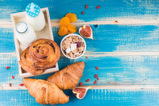 Pane al forno con bottiglie di latte nel vassoio vicino alle albicocche secche; frutto di fico; e corn flakes su sfondo blu in legno