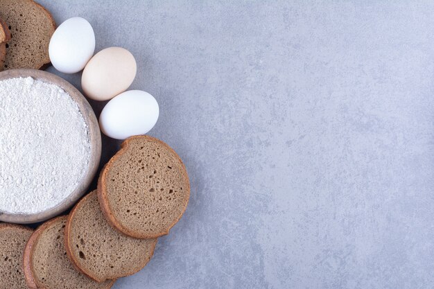 Pane nero a fette e uova intorno a una ciotola di farina sulla superficie di marmo