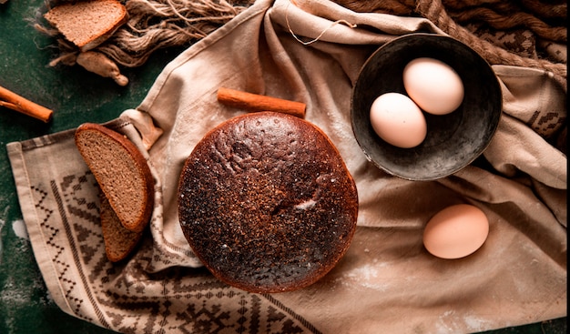 Pane nero rotondo con fette, cannella e ciotola di uova su un tavolo verde.