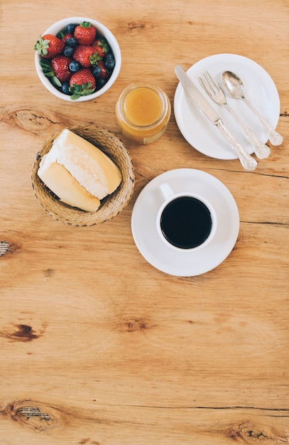 Pane; tazza di caffè; marmellata; bacche fresche e posate sul piatto contro sfondo in legno