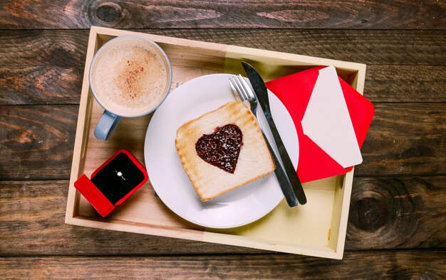 Pane tostato con marmellata sul piatto vicino a posate, tazza di bevanda, busta e anello in confezione regalo a bordo