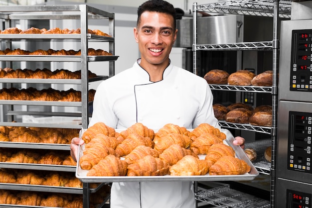 Panettiere che sorride alla macchina fotografica che tiene vassoio di croissant in una cucina commerciale