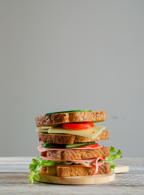 Panino con il pomodoro, il cetriolo, il formaggio, la salsiccia, verdi su un tagliere sulla tavola di legno e grigia, vista laterale.