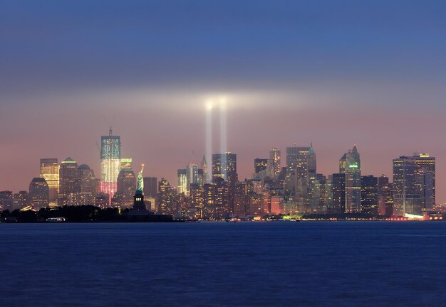 Panorama dello skyline del centro di New York City Manhattan di notte con la statua della libertà e fasci di luce in memoria dell'11 settembre visto dal lungomare del New Jersey.