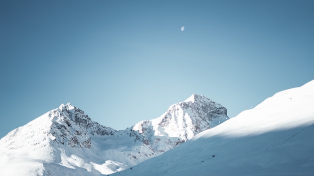 Panoramica delle montagne coperte di neve sotto un cielo blu chiaro con una mezza luna