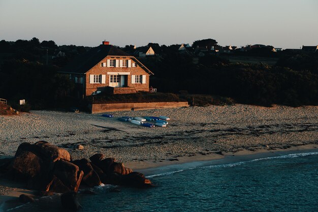 Panoramica di una casa marrone su una spiaggia sabbiosa vicino al mare circondata da rocce e alberi