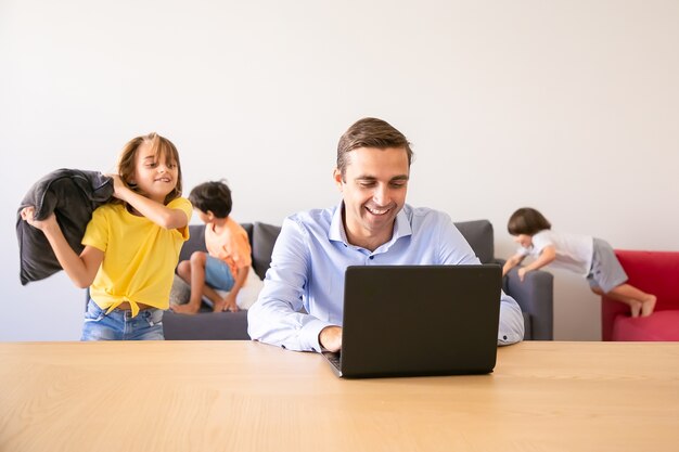 Papà allegro che chiacchiera tramite laptop e bambini che giocano con i cuscini vicino a lui. Padre caucasico che lavora a casa durante le vacanze scolastiche. Famiglia e concetto di tecnologia digitale