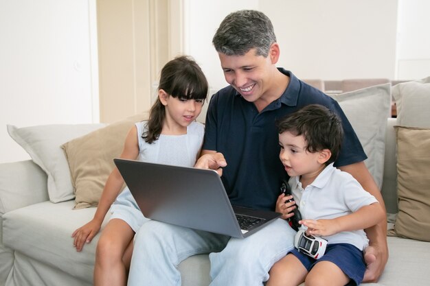 Papà allegro che mostra il contenuto sul laptop a due bambini curiosi. Famiglia che guarda film a casa.