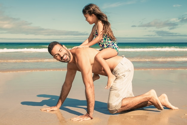 Papà allegro che va sulle mani e sulle ginocchia sulla spiaggia, portando la bambina sulla schiena