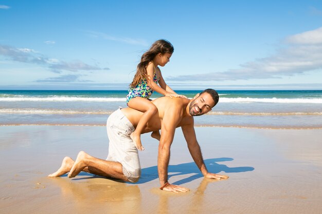 Papà allegro che va sulle mani e sulle ginocchia sulla spiaggia, portando la bambina sulla schiena