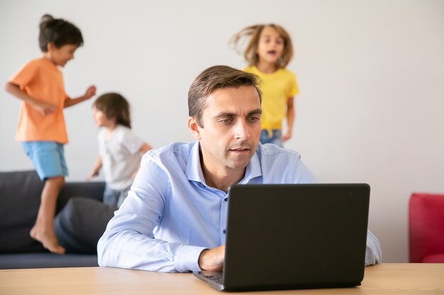 Papà caucasico serio che lavora tramite laptop e bambini che saltano. Padre concentrato utilizzando computer e bambini che giocano sul divano. Messa a fuoco selettiva. Infanzia e concetto di tecnologia digitale