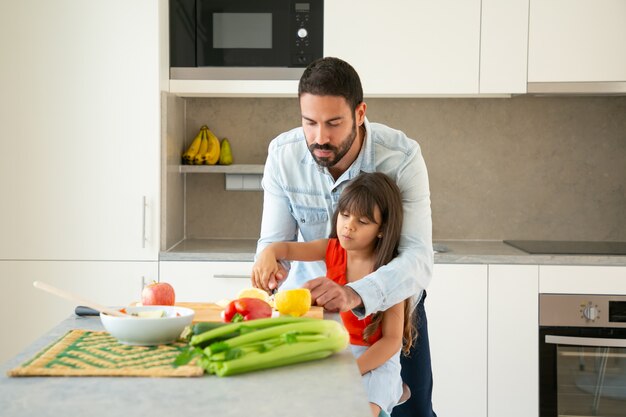 Papà che insegna alla figlia a preparare l'insalata. Ragazza e suo padre che tagliano la verdura fresca al bancone della cucina. Concetto di cucina familiare