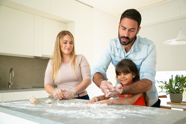 Papà concentrato che insegna alla figlia a fare la pasta sul tavolo della cucina con farina disordinata. I giovani genitori e la loro ragazza cuociono insieme focacce o torte. Concetto di cucina familiare