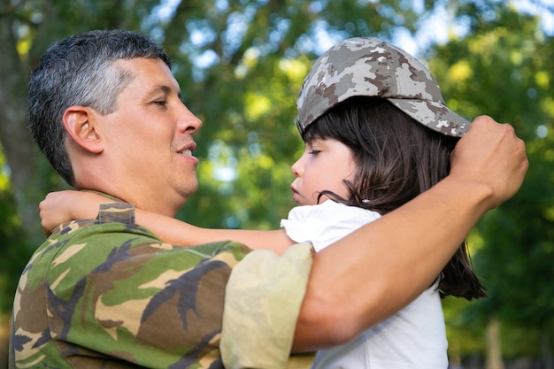 Papà felice che tiene la figlia tra le braccia, vestendo la ragazza nel suo berretto mimetico. Vista laterale. Ricongiungimento familiare o concetto di ritorno a casa