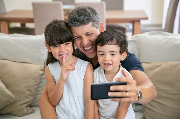 Papà felice e due bambini che prendono selfie o utilizzano il telefono per la videochiamata mentre erano seduti sul divano a casa insieme.