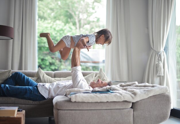 Papà sdraiato sul divano e solleva il bambino in aria sotto la luce del sole attraverso le finestre