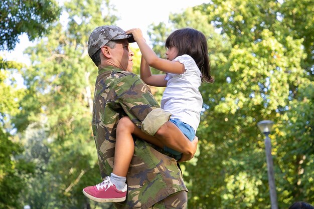 Papà serio in uniforme militare che tiene la figlia sulle mani, guardandola e in piedi all'aperto. Piccola ragazza sveglia concentrata che tocca la protezione del padre. Ricongiungimento familiare, paternità e concetto di fine settimana