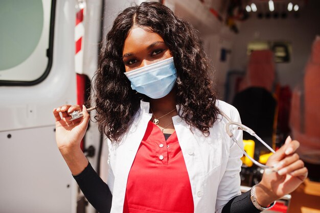 Paramedico femminile afroamericano in maschera medica protettiva per il viso in piedi davanti all'auto ambulanza