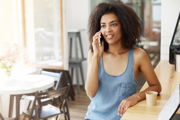 Parlare sorridente della bella donna africana allegra felice sul telefono che si siede in caffè.