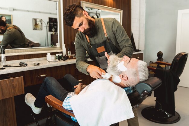 Parrucchiere che mette crema da barba al cliente anziano nel salone di bellezza