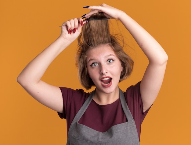 Parrucchiere di giovane bella donna in grembiule che pettina i suoi capelli che sembra felice e sorpreso in piedi sopra la parete arancione