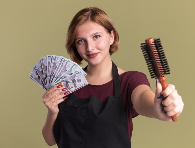 Parrucchiere di giovane bella donna in grembiule che tiene contanti che mostra la spazzola per capelli sorridente fiducioso in piedi sopra la parete verde