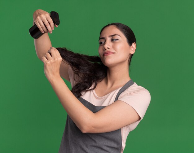 Parrucchiere di giovane bella donna in grembiule che tiene l'applicazione di spray idratante ai suoi capelli guardando fiducioso in piedi sopra la parete verde
