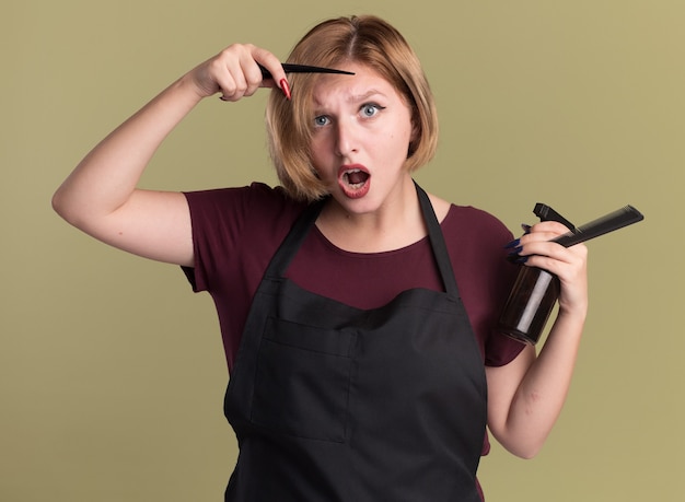 Parrucchiere di giovane bella donna in grembiule che tiene la bottiglia spray per capelli e pettine guardando davanti sorpreso in piedi sopra la parete verde