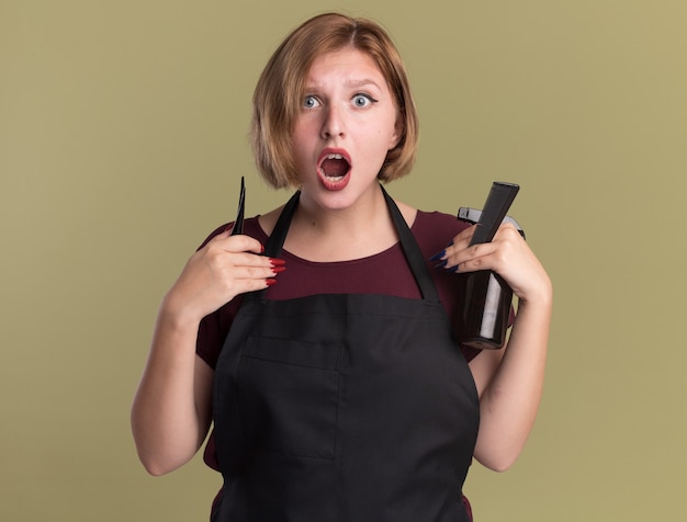 Parrucchiere di giovane bella donna in grembiule che tiene la bottiglia spray per capelli e pettine guardando davanti sorpreso in piedi sopra la parete verde