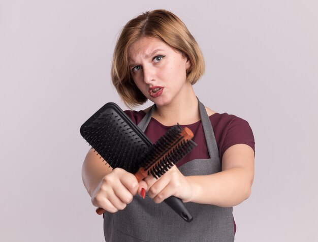 Parrucchiere di giovane bella donna in grembiule che tiene le spazzole per capelli che attraversano le mani guardando la parte anteriore con la faccia seria in piedi sopra il muro bianco