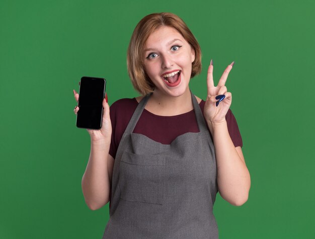 Parrucchiere di giovane bella donna in grembiule che tiene smartphone che mostra v-segno sorridente allegramente in piedi sopra la parete verde