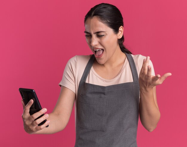 Parrucchiere di giovane bella donna in grembiule che tiene smartphone guardando che è confuso e deluso in piedi sopra la parete rosa