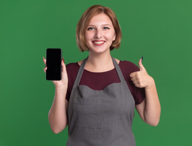 Parrucchiere di giovane bella donna in grembiule che tiene smartphone guardando davanti sorridendo allegramente mostrando i pollici in su in piedi sopra la parete verde