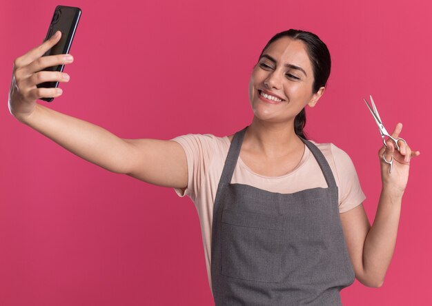 Parrucchiere di giovane bella donna in grembiule che tiene smartphone guardando facendo selfie mostrando le forbici sorridente in piedi sopra la parete rosa