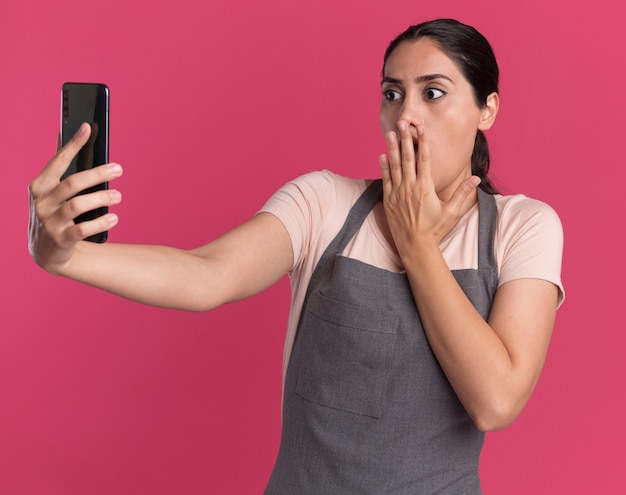 Parrucchiere di giovane bella donna in grembiule che tiene smartphone guardandolo stupito e sorpreso in piedi sopra il muro rosa