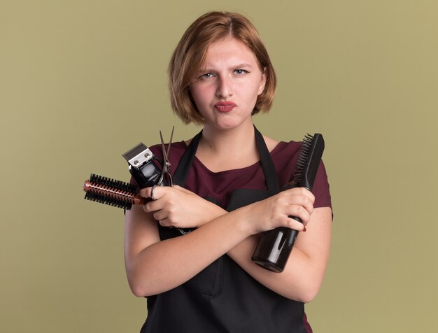 Parrucchiere di giovane bella donna in grembiule che tiene trimmer con spazzola per capelli e flacone spray guardando la parte anteriore dispiaciuto con le braccia incrociate in piedi sopra la parete verde