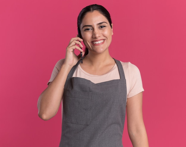 Parrucchiere di giovane bella donna in grembiule guardando davanti sorridente con la faccia felice mentre parla al telefono cellulare in piedi sopra il muro rosa