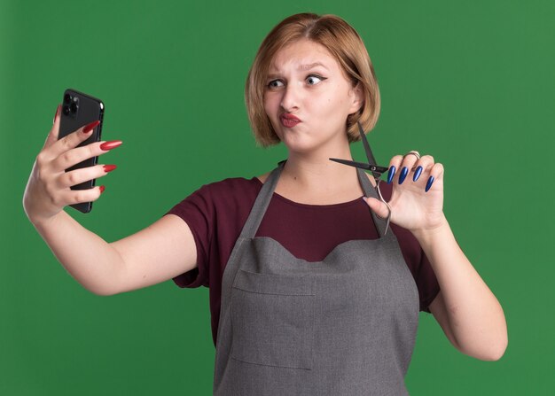 Parrucchiere di giovane bella donna in grembiule utilizzando smartphone facendo selfie con le forbici essendo confuso in piedi sopra la parete verde