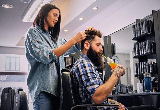 Parrucchiere femminile che taglia i capelli del maschio barbuto dei pantaloni a vita bassa in un salone.