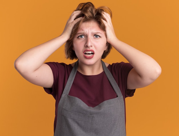 Parrucchiere giovane bella donna in grembiule frustrato tirando i capelli in piedi sopra la parete arancione