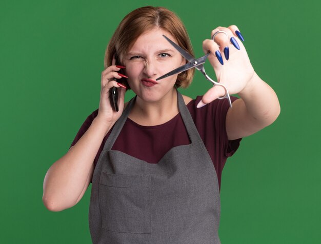 Parrucchiere giovane bella donna in grembiule parlando al telefono cellulare che mostra le forbici davanti guardando confuso e scontento in piedi sopra la parete verde